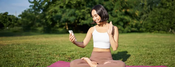 Portrait of asian girl say hello on smartphone, wave at mobile phone while doing meditation, online yoga classes on fresh air in park.