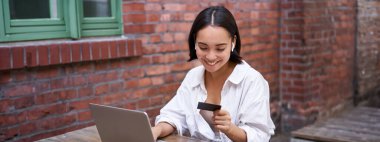 Bilgisayarlı ve kablosuz kulaklıklı gülümseyen Asyalı kadın, kredi kartıyla ödeme yapıyor, internetten alıyor, bir fincan kahveyle oturuyor..