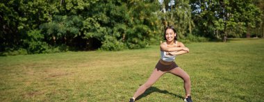 Genç Asyalı fitness kızı geriniyor, parkta egzersiz yapıyor, egzersiz yapıyor, çömelme hareketleri yapıyor..