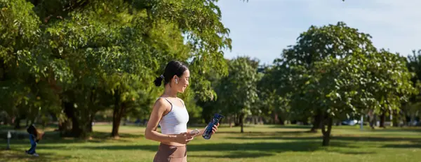 stock image Active asian girl, in fitness clothing, workout in park, walking in sportswear with smartphone and water bottle.