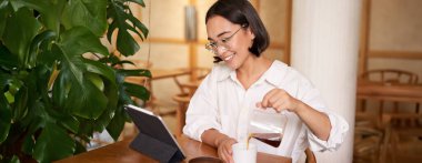 Serbest ve uzak işçiler. Gülümseyen genç bir kadın bir fincana kahve dolduruyor, kafede oturuyor ve dijital tablete bakıyor..