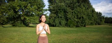 Dikey çekimde gülümseyen Asyalı kız yoga yapıyor, parkta egzersiz yapıyor, temiz havada lastik minderin üzerinde duruyor..