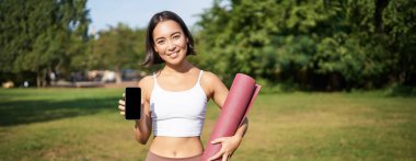 Kauçuk yoga minderiyle gülümseyen Asyalı fitness kızı, akıllı telefon ekranını gösteriyor, egzersiz uygulaması öneriyor, parkta çimlerde duruyor..