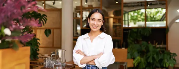 Young asian woman, cafe owner or manager, standing confident with arms crossed on hands, smiling and looking at camera.