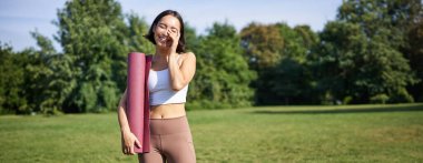 Asyalı kız portresi gülümsüyor ve gülüyor, spor malzemeleriyle ayakta duruyor, lastik paspas, spor için üniforma giyiyor ve parkta egzersiz yapıyor..