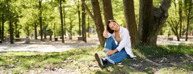 Rahatlamış genç bir kadın, ağacın yanında dinleniyor, parkta gölgenin altında oturuyor, gülümsüyor ve mutlu görünüyor, açık havada temiz havada yürüyor..