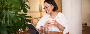 Gözlüklü Asyalı kadın, dijital tabletle smth izliyor, kafede kahve içiyor ve gülümsüyor, uzaktan çalışıyor..