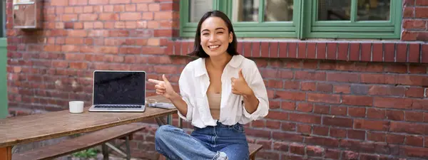 stock image Stylish young asian woman sitting in cafe with laptop, outdoor table, pointing finger at advertisement, showing banner copy space.