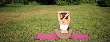 Sporcunun antrenmanın ortasında arka tarafa bakması, amrs arkasını germesi, parkta çimenlikte kauçuk yoga minderinde oturması..