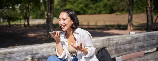 Portrait of happy asian girl records voice message, talking via speakerphone on smartphone, sitting relaxed on bench in park on sunny day.