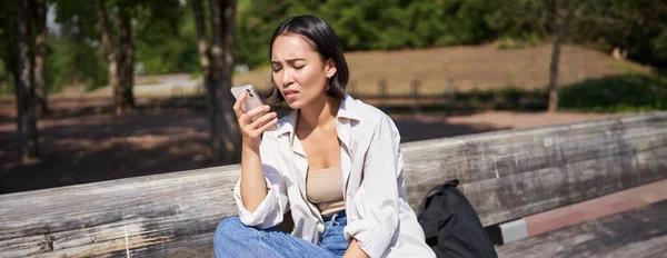 Asian girl frowning, looking concerned at her mobile phone, reading sad message, bad news on smartphone app, sitting on bench in park worrying.