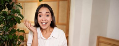 Şaşırmış görünen Asyalı bir kızın portresi, elinde şaşkın bir yüzle cep telefonu, akıllı telefondan harika haberler, kitapla kafede oturuyor..