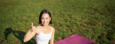 Parktaki yoga eğitiminden sonra kauçuk paspasın üzerinde oturan Asyalı bir kadının dikey çekimi onaylanmış başparmağını gösteriyor. Temiz hava üzerine meditasyon..