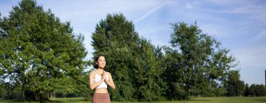 Genç, düşünceli bir kadın, temiz hava üzerine meditasyon yapıyor, parkta yoga egzersizleri yapıyor, spor kıyafetleri içinde lastik minderin üzerinde duruyor..