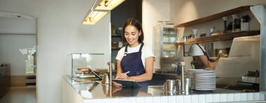 Gülümseyen güzel Asyalı bir kızın portresi, tezgahın arkasında çalışan bir barista, POS terminali olarak tablet kullanımı, işlem düzeni..