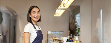 Dikey olarak gülümseyen Asyalı kız barista, üniforma giyiyor, kahve yapıyor, fincanla tezgahın yanında duruyor, kafede çalışıyor..