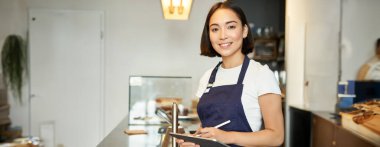 Küçük kafe işi. Önlüklü gülümseyen Asyalı kız, POS terminali olarak tableti kullanıyor, tezgahın arkasında kahve siparişini işliyor..