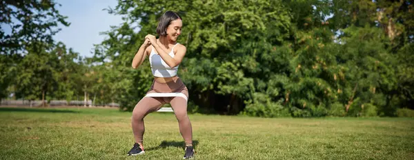 Woman using stretching band, resistance fitness rope for workout in park, doing squats, training on fresh air.