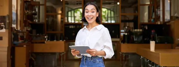 Smiling beautiful waitress, businesswoman with digital tablet, laughing and looking happy, standing in front of cafe or restaurant.