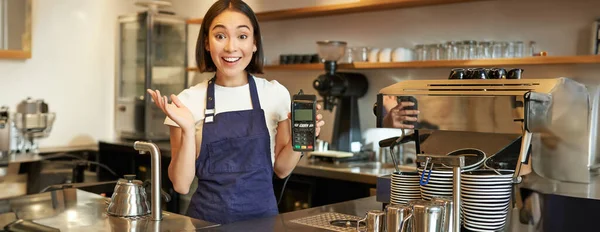 Enthusiastic asian girl barista in apron, showing contactless credit card machine, looks amazed by fast contactless payment, working in coffee shop.