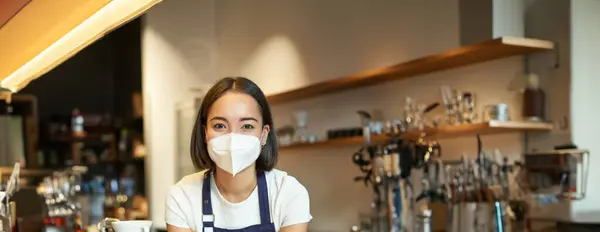 Young female barista in apron and medical mask, working in cafe during covid, leaning on counter and looking at camera, taking orders from guests, making coffee.