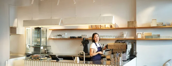 Portrait of cafe counter with barista girl working with coffee machine, making order for client in shop, wearing blue apron.