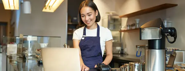 Portrait of young girl cafe owner, looking at her laptop, taking order, serving customer in coffee shop.