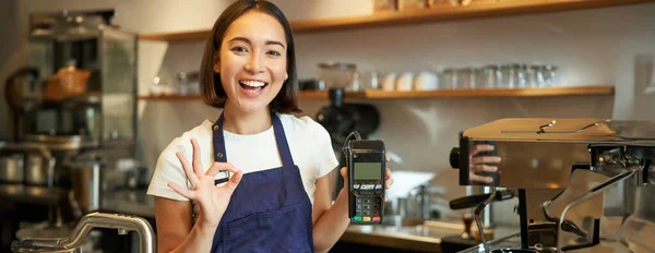 Smiling asian barista girl, wears apron, shows credit card machine for processing payment, suggest to pay contactless, standing in coffee shop.