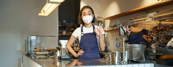 Asian female barista, girl in medical mask, shows okay sign, works in cafe behind counter, brews coffee, works with clients during covid pandemic.