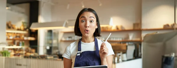 Enthusiastic asian female entrepreneur, cafe owner with tablet, wearing shop uniform, standing with tablet and pen and looking excited.