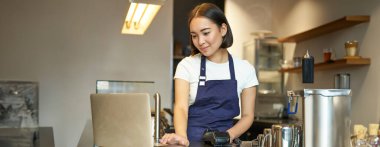 Gülümseyen Asyalı kız barista, kafede çalışıyor, müşteriye hizmet ediyor, dizüstü bilgisayara bakıyor ve sipariş için para alıyor, kahve satıyor..