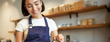 Esmer Asyalı baristanın dikey pozu, önlük giyiyor, kahve yapıyor, kafede sipariş hazırlıyor, kapuçino latte sanatı için sütü bardağa dolduruyor..