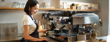 Mutlu Asyalı kız, önlüklü barista kahve makinesiyle tezgahın arkasında çalışıyor, kafede duruyor ve cappuccino yapıyor..