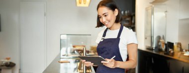 Küçük kafe işi. Önlüklü gülümseyen Asyalı kız, POS terminali olarak tableti kullanıyor, tezgahın arkasında kahve siparişini işliyor..