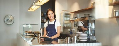 Gülümseyen güzel Asyalı bir kızın portresi, tezgahın arkasında çalışan bir barista, POS terminali olarak tablet kullanımı, işlem düzeni..