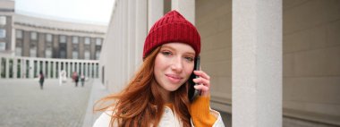 Mobile broadband and people. Smiling young redhead woman walks in town and talks on mobile phone, calling friend on smartphone, using internet to make a call abroad.