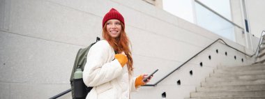 Redhead girl, young woman tourist with backpack, holds smartphone, looks for route on mobile application, searches for hotel on phone map, smiles happily.