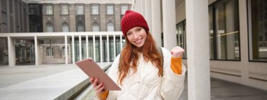 Stylish ginger girl, tourist walks with digital tablet around city, woman connects to iternet on her gadget, looking up information, texting message.