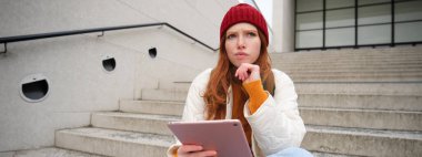 Portrait of troubled redhead girl, college student with thoughtful face, sits on street stairs, holds digital tablet, thinks how to reply on message.