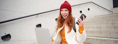 Smiling redhead woman with mobile phone and laptop, sitting on stairs outside building, connects to public wifi, using smartphone and computer.