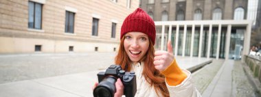 Beautiful readhead girl, photographer with professional camera takes pictures outdoors, walking around city and taking photos, sightseeing.