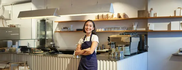 Portrait of cute asian woman barista, cafe staff standing near counter with coffee machine, wearing apron, smiling at camera.