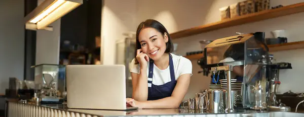 Portrait of smiling korean woman, barista in coffee shop, standing at counter with laptop, smiling and looking confident, self-employed female entrepreneur in her own coffee shop.