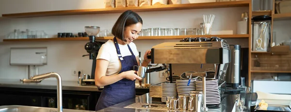 Happy asian girl barista, working in cafe, brewing coffee, prepare cappuccino, using steamer on machine to prepare latte, smiling and laughing.