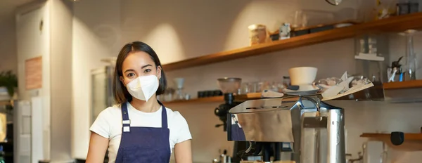 Vertical shot of friendly barista, female staff member in medical mask, working behind counter with clients, serving coffee in cafe, standing in uniform apron.