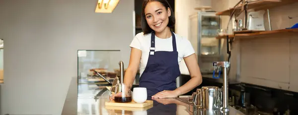 Smiling beautiful barista girl in apron, making batch brew, filter coffee, standing in cafe behind counter.