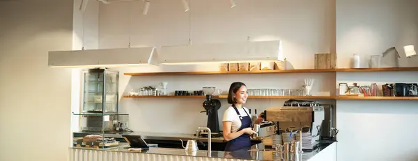 Portrait of cafe counter with barista girl working with coffee machine, making order for client in shop, wearing blue apron.