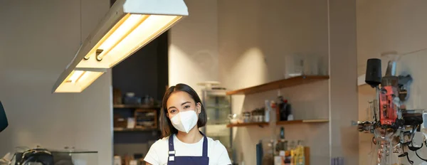 Vertical shot of friendly barista, female staff member in medical mask, working behind counter with clients, serving coffee in cafe, standing in uniform apron.