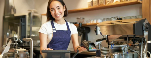 Small business and people. Smiling asian woman barista, working in cafe, giving POS terminal, credit card machine, processing order.