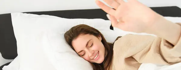 Portrait of gorgeous girlfriend stretching hand, blocking camera, lying in bed and resting on pillow, smiling while waking up from someone.